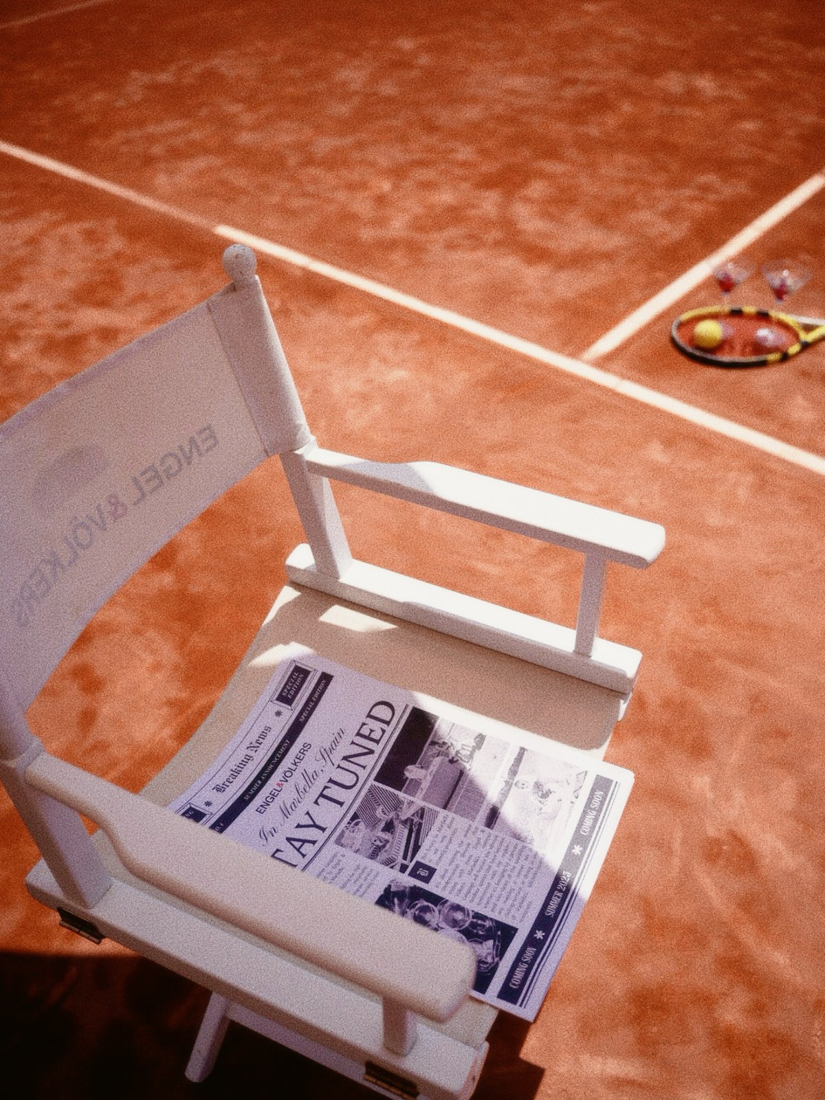 A custom "Stay Tuned" newspaper resting on an Engel & Volkers director's chair on a clay tennis court
