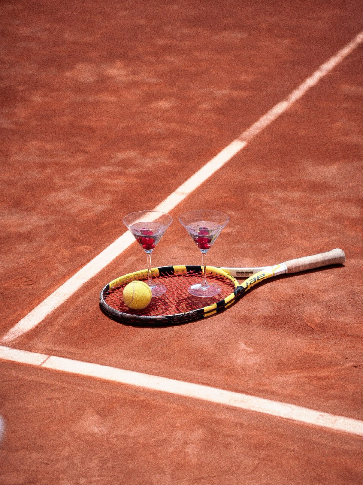 A tennis racket holding two martini glasses and a ball on a sunlit clay court
