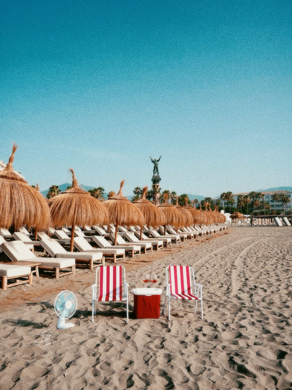 A retro Marbella beach scene with striped deck chairs, a cool box and a fan on the sand