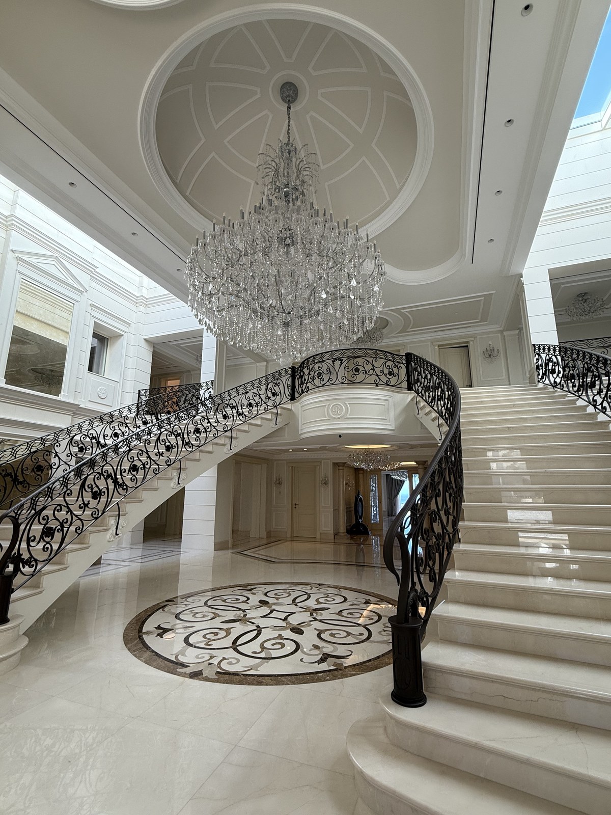 The villa's grand marble entrance hall, with a sweeping wrought-iron staircase and a crystal chandelier beneath a domed ceiling