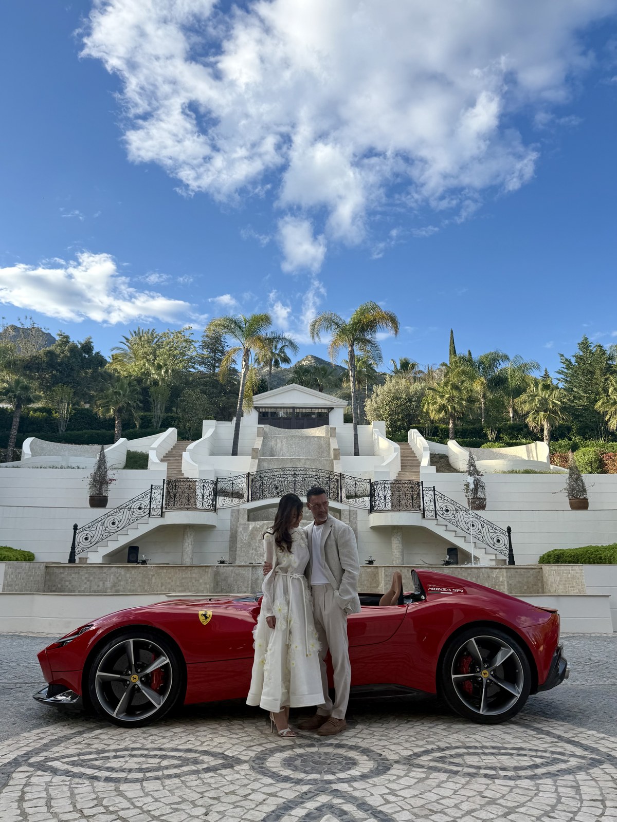 A couple beside a red Ferrari Monza SP1 in front of the grand white staircase of a luxury Marbella villa